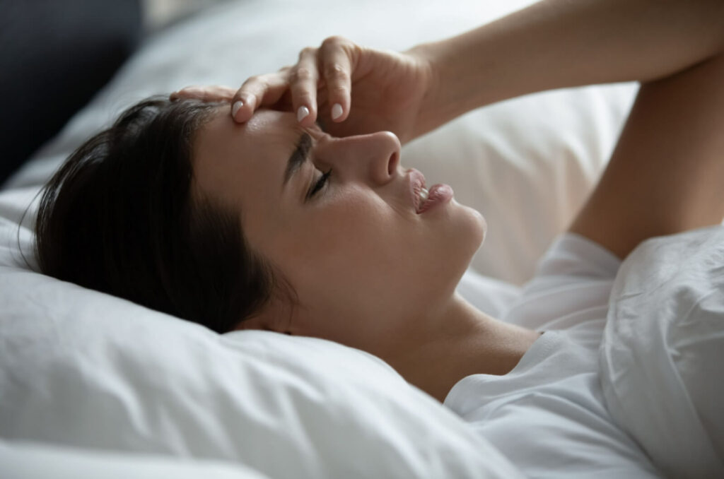 A woman lying in bed rubbing her forehead in frustration due to waking up with blurry eyesight.