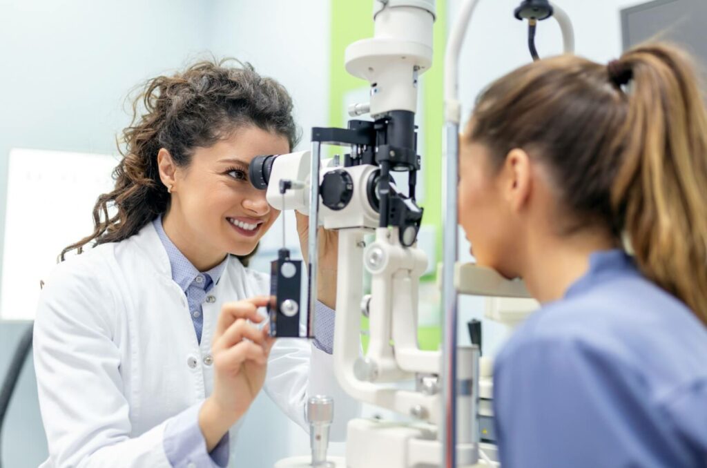 An optometrist smiling while examining her patient's eyes to determine why her eyesight is blurry when she wakes up.