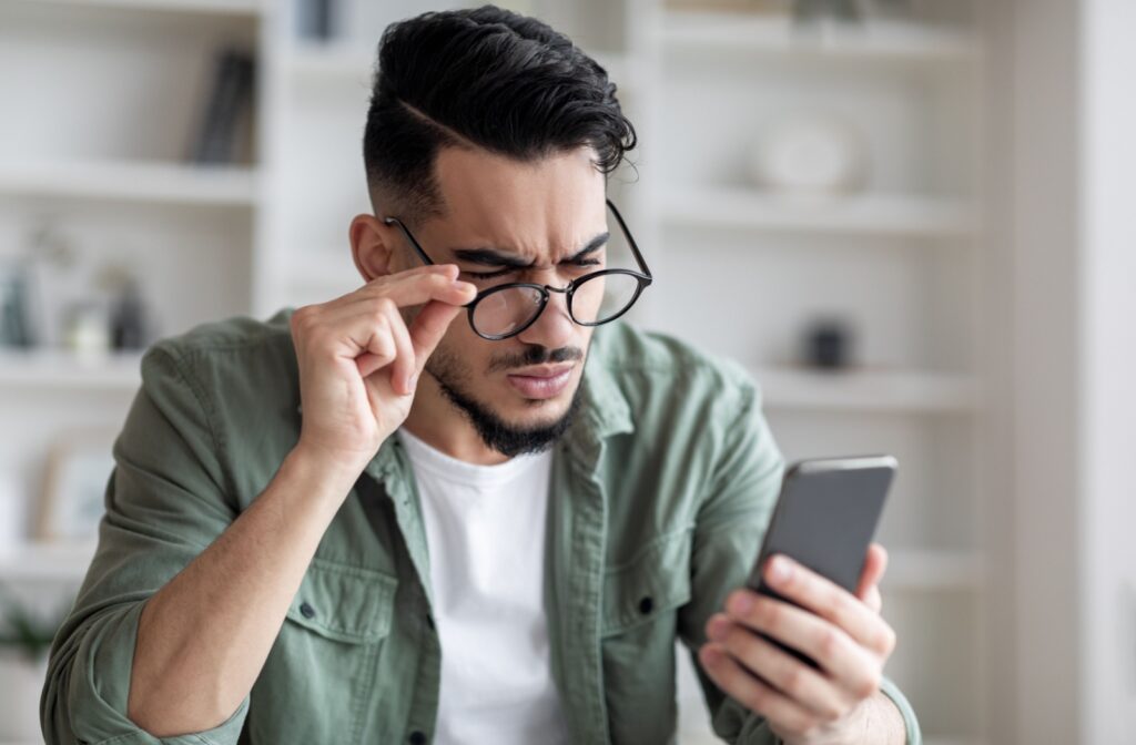 A person holds a smartphone in their left hand, sliding their glasses down their nose to see the screen more clearly, likely due to myopia and astigmatism.