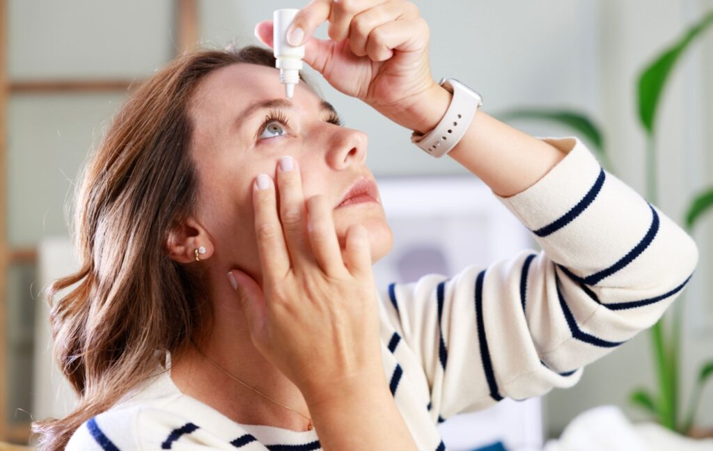 A person wearing a striped shirt looks up as they gently squeeze eye drops into their eye.