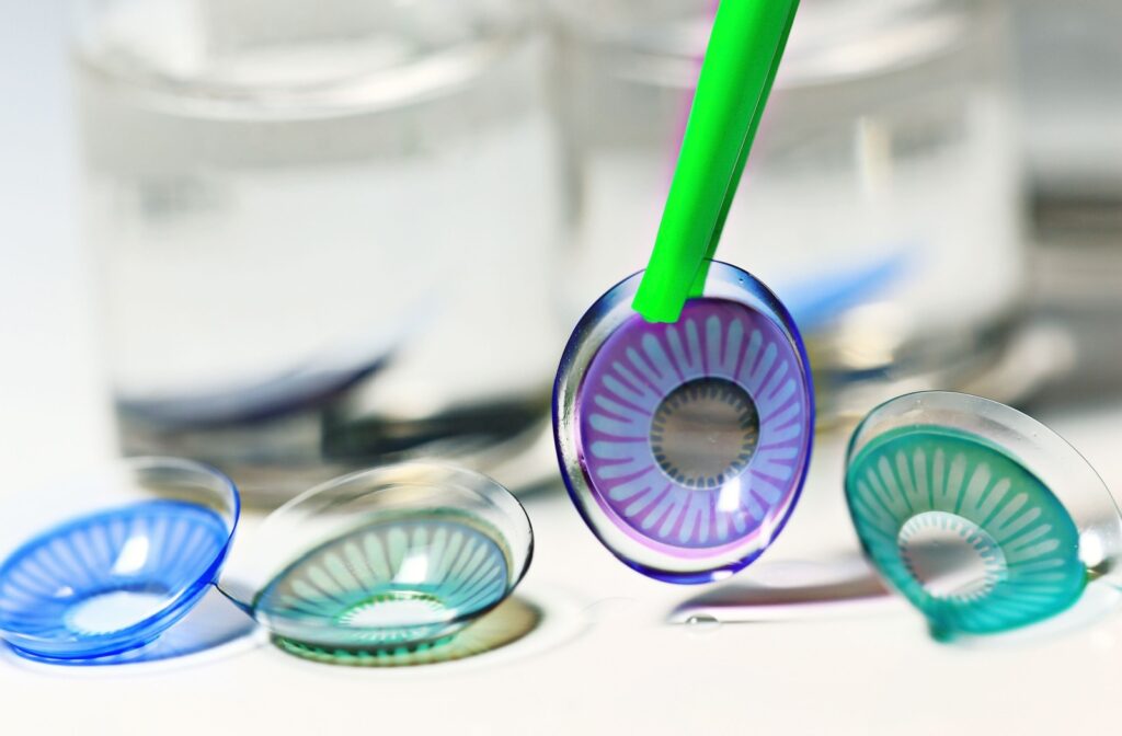 Several colorful contact lenses on a white surface with one being picked up by green tweezers.